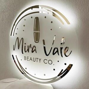 May include: A round, illuminated sign with the text "Mira Vale BEAUTY CO." in a stylish font. The sign features a lipstick graphic and a decorative border. The sign is white with a gold border and is mounted on a white wall.
