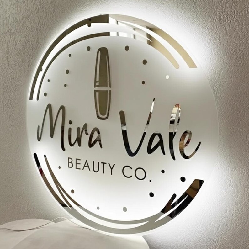 May include: A round, illuminated sign with the text "Mira Vale BEAUTY CO." in a stylish font. The sign features a lipstick graphic and a decorative border. The sign is white with a gold border and is mounted on a white wall.