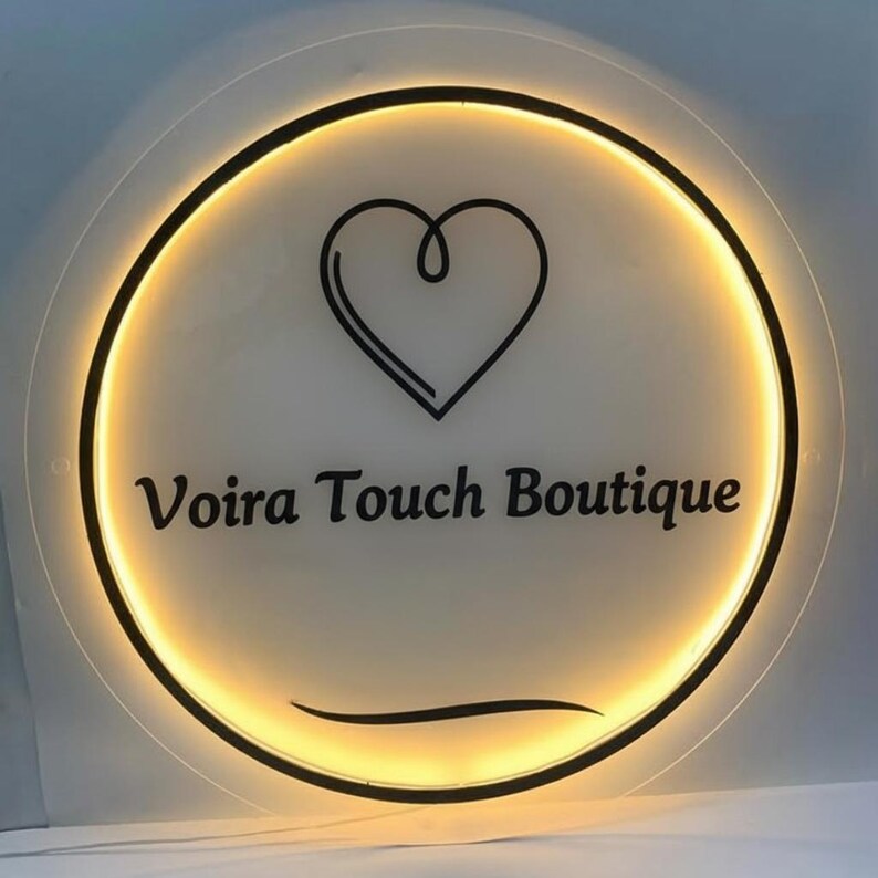 May include: Circular sign with the text "Voira Touch Boutique" in black, centered below a black heart outline. The sign has a warm, yellow glow emanating from within a black border.