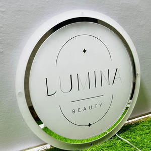 K&ouml;nnte beinhalten: Ein rundes, beleuchtetes Schild mit dem Text "LUMINA BEAUTY" in Schwarz. Das Schild hat einen wei&szlig;en Rahmen mit einem silbernen Innenring. Der Hintergrund ist eine hellgr&uuml;ne Oberfl&auml;che.