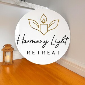 May include: A white, circular sign with the words "Harmony Light Retreat" in black script. The sign features a gold-colored candle and leaf design. A wooden lantern with a lit candle sits on the wooden floor.