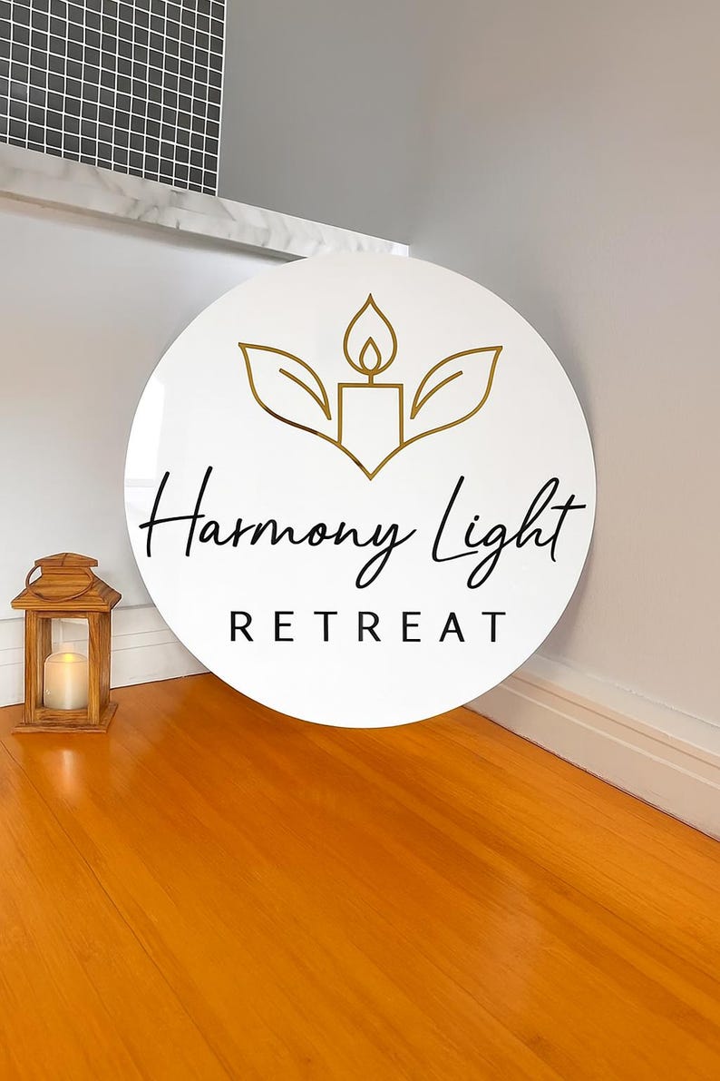 May include: A white, circular sign with the words "Harmony Light Retreat" in black script. The sign features a gold-colored candle and leaf design. A wooden lantern with a lit candle sits on the wooden floor.