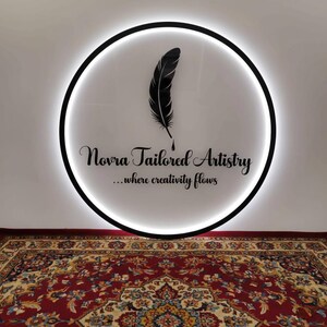 May include: A circular sign with a black border and a white illuminated interior. The sign features a black feather design and the text "Novra Tailored Artistry ...where creativity flows". A patterned rug is in the foreground.