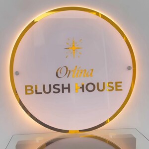 May include: A round sign with a gold border and the words "Orlina BLUSH HOUSE" in gold lettering. A gold starburst design is above the word "Orlina". The sign is mounted on a clear glass surface.