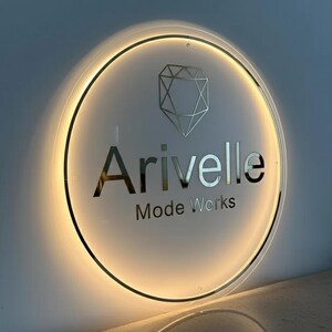 May include: A backlit, circular sign with the name "Arivelle" in silver lettering, and the words "Mode Works" below. A geometric diamond shape is above the name. The sign is illuminated with a warm, golden light.