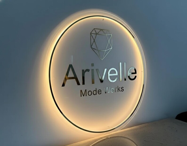 May include: A backlit, circular sign with the name "Arivelle" in silver lettering, and the words "Mode Works" below. A geometric diamond shape is above the name. The sign is illuminated with a warm, golden light.