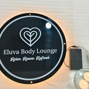 May include: A round, black sign with white text that reads "Eluva Body Lounge" and "Relax, Renew, Refresh." The sign has a white heart-shaped logo and is illuminated with a warm, soft light. A battery pack and small candles are also visible.
