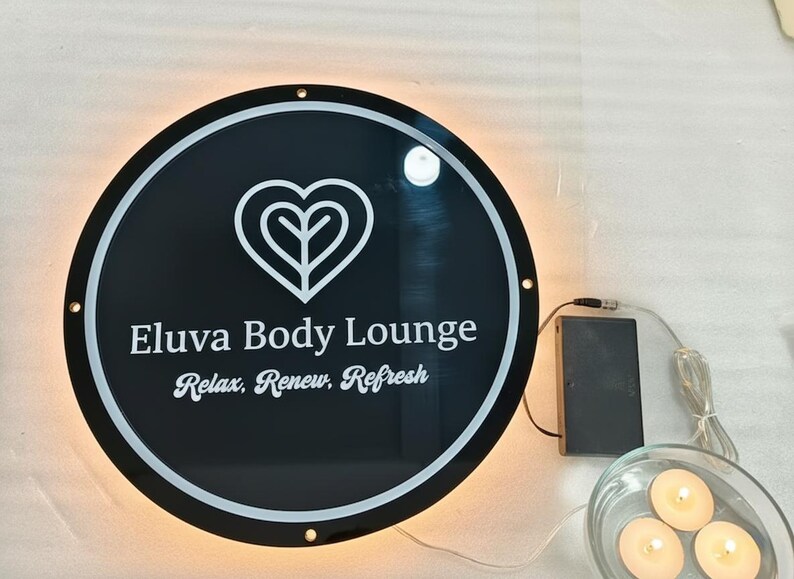 May include: A round, black sign with white text that reads "Eluva Body Lounge" and "Relax, Renew, Refresh." The sign has a white heart-shaped logo and is illuminated with a warm, soft light. A battery pack and small candles are also visible.