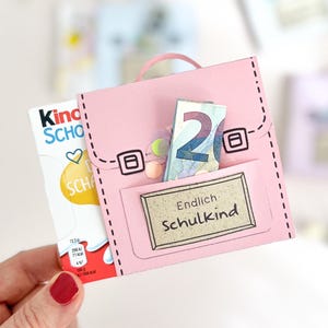 Può includere: Un lavoretto in carta rosa a forma di piccolo zaino con manico e tasca frontale. La tasca presenta un'etichetta con il testo Endlich Schulkind. Una barretta di cioccolato Kinder e una banconota da venti euro piegata sono inserite nello zaino.