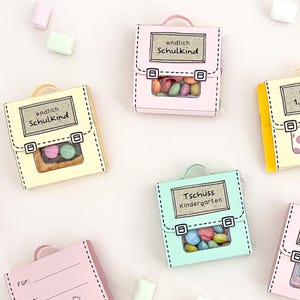 Könnte beinhalten: Eine Sammlung kleiner, bunter Geschenkschachteln aus Papier in Form von Schulranzen in Pastelltönen wie Rosa, Gelb und Mintgrün. Jede Schachtel hat einen Griff, ein Sichtfenster mit kleinen Süßigkeiten und Etiketten mit Text wie endlich SCHULKIND.