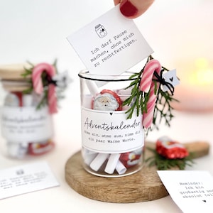 Könnte beinhalten: Ein durchsichtiges Glas gefüllt mit gerollten Zetteln und Schokoladen-Weihnachtsmännern, mit einem Etikett mit der Aufschrift Adventskalender. Das Glas ist mit einer Zuckerstange und einem Tannenzweig verziert. Ein Zettel darüber zeigt den Text Ich darf Pause machen, ohne mich zu rechtfertigen.