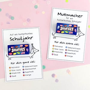 以下が含まれることがあります： パーティーハットをかぶったダックスフントの黒い線画が描かれた、2枚の白い長方形のカード。各カードには、小さな青い箱に入ったSmarties Miniキャンディが取り付けられています。テキストには「Auf ein kunterbuntes Schuljahr」および「Mutmacher」と書かれています。