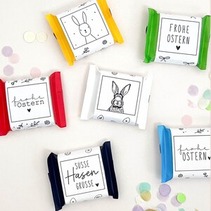 Könnte beinhalten: Eine Auswahl an Oster-Schokoriegel-Verpackungen in verschiedenen Farben, darunter Gelb, Grün, Rot, Weiß, Blau und Marineblau. Jede Verpackung zeigt ein anderes Design, wie eine Hasenillustration oder den Text "Frohe Ostern" und "Süsse Hasen Grüsse".