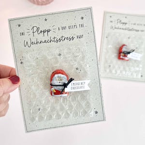 May include: A light grey greeting card featuring a square of clear bubble wrap and a small chocolate Santa Claus figure tied with black string. The card displays the text One Plopp a day keeps the Weihnachtsstress away and Emergency Chocolate.