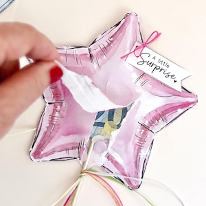 Könnte beinhalten: Ein sternförmiger Papierballon in Rosatönen mit Aquarell-Effekt, versehen mit einem weißen Anhänger mit dem Text A little Surprise, befestigt an einer rosa Schnur. Der Ballon hat einen Holzstab an der Basis mit bunten Bändern.