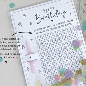 May include: A birthday card with a word search puzzle, confetti, and two pink candles. The card reads "Happy Birthday" in black script. The word search has German words. The card is in a clear plastic sleeve.