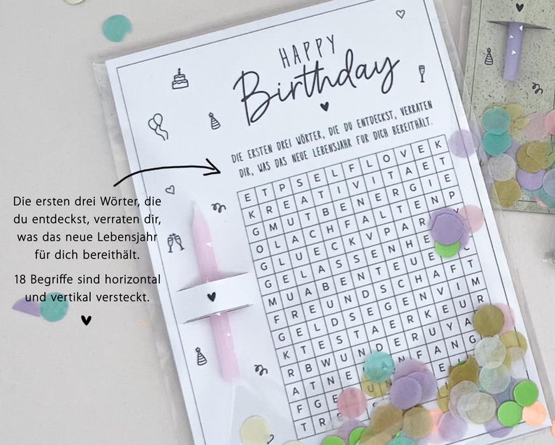 May include: A birthday card with a word search puzzle, confetti, and two pink candles. The card reads "Happy Birthday" in black script. The word search has German words. The card is in a clear plastic sleeve.