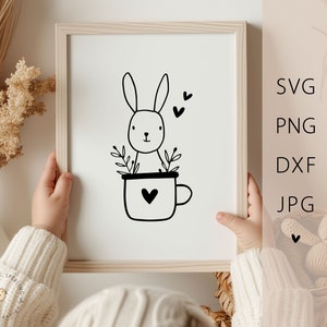 May include: Black and white illustration of a bunny rabbit sitting in a coffee cup with a heart on the side. The bunny has small ears and a heart above its head.