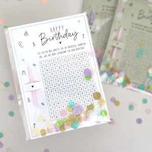 May include: A clear plastic bag containing a birthday-themed word search puzzle with the words "HAPPY Birthday" at the top. The bag also contains confetti and a pink candle. The background is white.