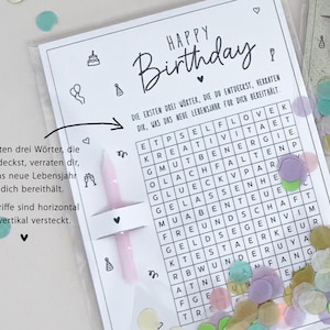 May include: A birthday card with a word search puzzle, confetti, and two pink candles. The card reads "Happy Birthday" in black script. The word search has German words. The card is in a clear plastic sleeve.
