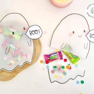 Könnte beinhalten: Zwei geisterförmige Papiertüten mit schwarzem Umriss, jeweils mit einer Sprechblase und dem Text BOO!. Eine Tüte ist mit bunten Bonbons und kleinen Pompons gefüllt, während die andere leicht geöffnet ist und Bonbons sowie Konfetti zeigt.