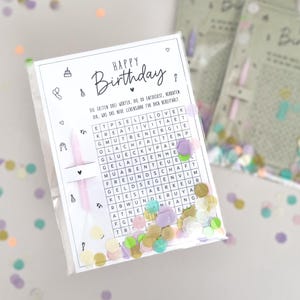May include: A clear cellophane bag filled with confetti and a white card with the words "Happy Birthday" and a word search puzzle. The card includes small illustrations and text in German. A pink candle is included.