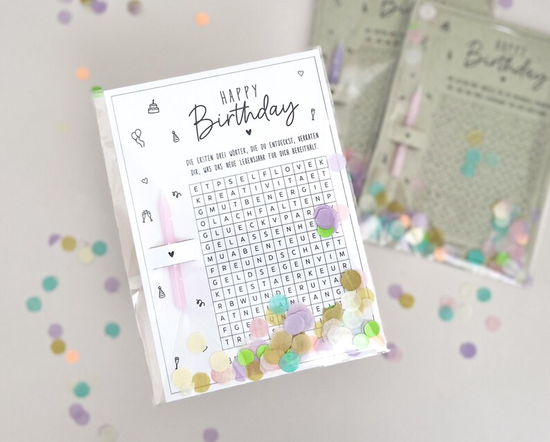 May include: A clear cellophane bag filled with confetti and a white card with the words "Happy Birthday" and a word search puzzle. The card includes small illustrations and text in German. A pink candle is included.