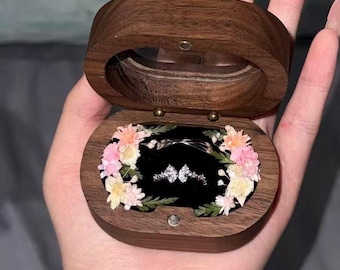May include: A wooden ring box with a clear lid, open to reveal a ring on a black velvet base. The box is decorated with a floral arrangement of pink, peach, and white flowers. The ring is silver with two diamond-shaped stones.