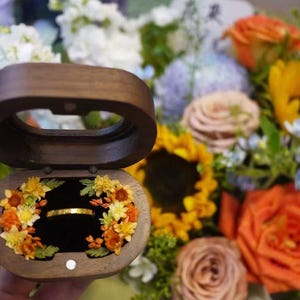 May include: A wooden ring box with a gold ring inside, surrounded by a floral arrangement of yellow and orange flowers. The box is open, revealing the ring against a black interior. A bouquet of flowers is in the background.