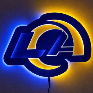 Może przedstawiać: Naścienny znak z logo Los Angeles Rams przedstawiający litery LA w kolorze ciemnoniebieskim oraz zakrzywiony kształt rogu. Znak jest podświetlony niebieską poświatą za literami i żółtą poświatą za rogiem, tworząc żywy efekt świetlny.