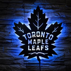 Può includere: Un'insegna da parete in metallo nero a forma di foglia d'acero con il testo TORONTO MAPLE LEAFS ritagliato al centro. L'insegna è illuminata da dietro con una luce LED blu brillante, creando un effetto luminoso attorno ai bordi della foglia.