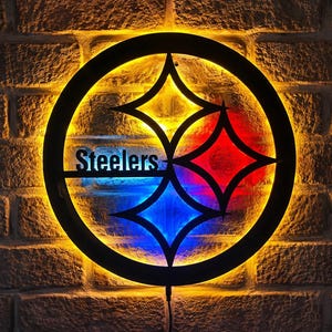 May include: A circular black metal wall sign featuring the Pittsburgh Steelers logo with the word Steelers in black lettering. The sign is illuminated from behind with a bright yellow glow, while the three diamond shapes are lit in yellow, red, and blue.