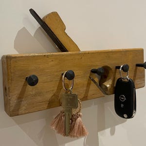 Könnte beinhalten: Ein Schlüsselhalter aus einem recycelten Handhobel aus Holz. Der Schlüsselhalter hat vier schwarze Metallstifte und hält Schlüssel mit rosa Quasten und einen Autoschlüssel. Das Holz ist hellbraun.