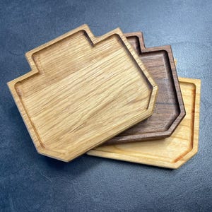 Könnte beinhalten: Drei Holztabletts in verschiedenen Brauntönen, übereinander gestapelt. Das obere Tablett ist aus hellem Eichenholz, das mittlere aus dunklerem Walnussholz und das untere aus hellerem Holz. Jedes Tablett hat eine einzigartige geometrische Form.