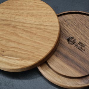 Könnte beinhalten: Zwei Untersetzer aus Holz, einer aus hellem Eichenholz und einer aus dunklem Walnussholz, sind abgebildet. Der dunkle Walnuss-Untersetzer trägt den Text "Holz Feder" und ein Logo. Die Untersetzer sind rund und haben eine natürliche Holzmaserung.