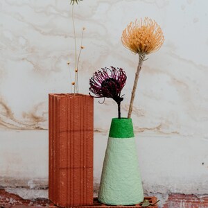May include: Dried flowers in a green and white cone-shaped vase made of paper mache. The vase is sitting on top of two red brick blocks.