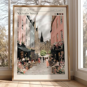 Puede incluir: Impresión enmarcada de Victoria Street en Edimburgo, Escocia. La obra de arte representa una escena callejera colorida con edificios rosas, azules y beige, cafés y personas. La impresión incluye el texto "Edinburgh, Scotland Victoria Street".