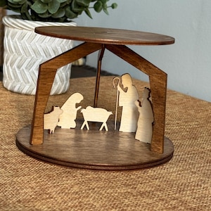 May include: A wooden nativity scene with a dark brown roof and base. The scene features cut-out figures of Mary, Joseph, the baby Jesus, and animals. The figures are light-colored, contrasting with the darker wood.