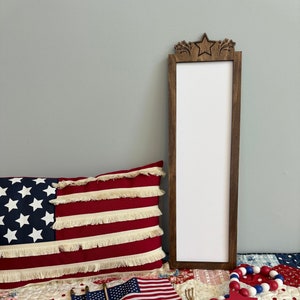 May include: A wooden sign with a star and fireworks design at the top. The sign is white and has the word "USA" written on it. The sign is sitting on a red, white, and blue quilt with a red, white, and blue beaded garland and a red star with the word "USA" written on it.