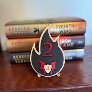 May include: A wooden, flame-shaped decorative item with a black centre and a light wood border. The number "2" and a shield with the number "4" are in red. The item is displayed in front of a stack of books.