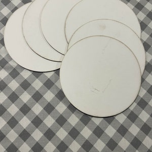 May include: A stack of white, round coasters with slightly aged edges. The coasters are arranged on a gray and white checkered pattern surface, creating a simple, clean aesthetic. The coasters are made of a sturdy material.