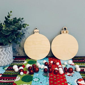 May include: Two unfinished wooden Christmas ornament cutouts with decorative tops. The ornaments are on a colorful quilted fabric with red, white, and green Christmas decorations. Small red and white ornaments are scattered around.