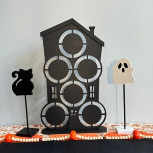 May include: Black wooden Halloween house with five cut-out circles, a black cat silhouette, and a white ghost silhouette. The house is on a table with a white bead garland and orange pumpkins.
