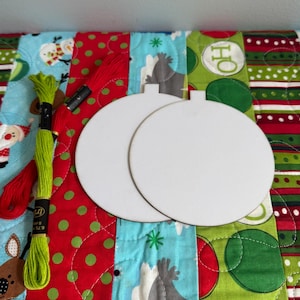 May include: Two blank, white, round ornaments with a hole at the top, ready for decoration, sit on a colorful Christmas-themed quilt. Green and red embroidery floss and a black bobbin are also present.