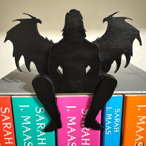 May include: Black wooden bookend shaped like a winged figure sitting on a stack of books with the author's name "J. Maas" and the title "Sarah" visible on the spines.