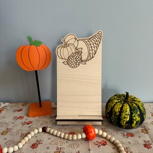 May include: A wooden sign with a fall harvest theme. The sign features a cornucopia with pumpkins, an apple, and a raspberry. The sign is on a stand and is ready to be used as a table top display.