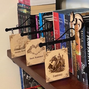 May include: Wooden book shelf signs with black metal hangers. The signs are engraved with the words "Steamy Read", "Dark Romance", and "Fantasy", along with related imagery. Books are visible in the background.