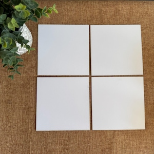 May include: Four blank, square white cards arranged in a grid pattern on a textured brown surface. A decorative plant with green leaves sits in the upper left corner. The cards are ready for custom designs.