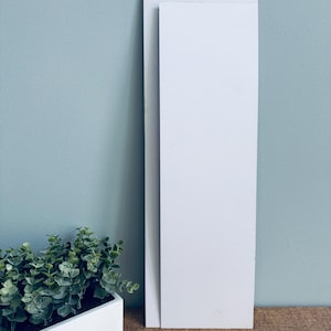 May include: Two white rectangular panels of different sizes are positioned vertically against a light blue wall. A rectangular planter with green plants is in the lower left corner. The panels are likely for home decor or DIY projects.