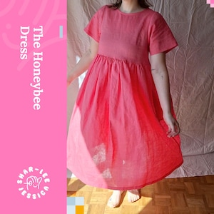 May include: A coral-coloured dress with short sleeves and a gathered waist. The dress features the text "The Honeybee Dress" and the designer's name. The dress is made of a lightweight fabric and has a flowing skirt.